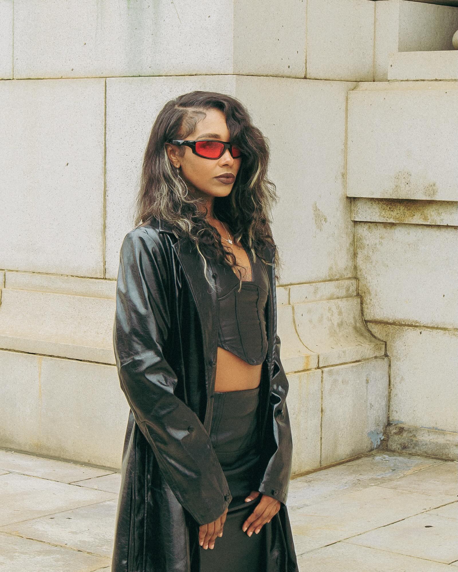 Featured portrait of Slycee in a black leather trench and red sunglasses against a stone wall